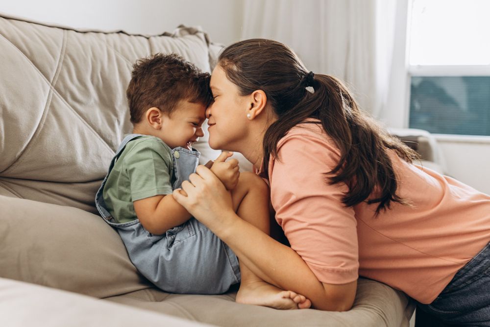 A mother and her toddler laughing forehead to forehead on the sofa